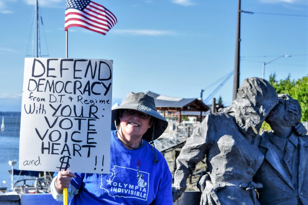 A member of Olympia Indivisible stands on the Olympia Boardwalk holding a sign saying 'Defend Democracy from D.T. + Regime with your: VOTE, VOICE, HEART, and $ !!'