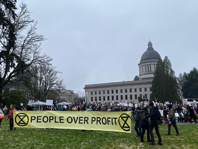 Protestors in front of the Olympia Capitol Building hold a large sign bearing the Extinction Rebellion logo and the phrase "People Over Profit"